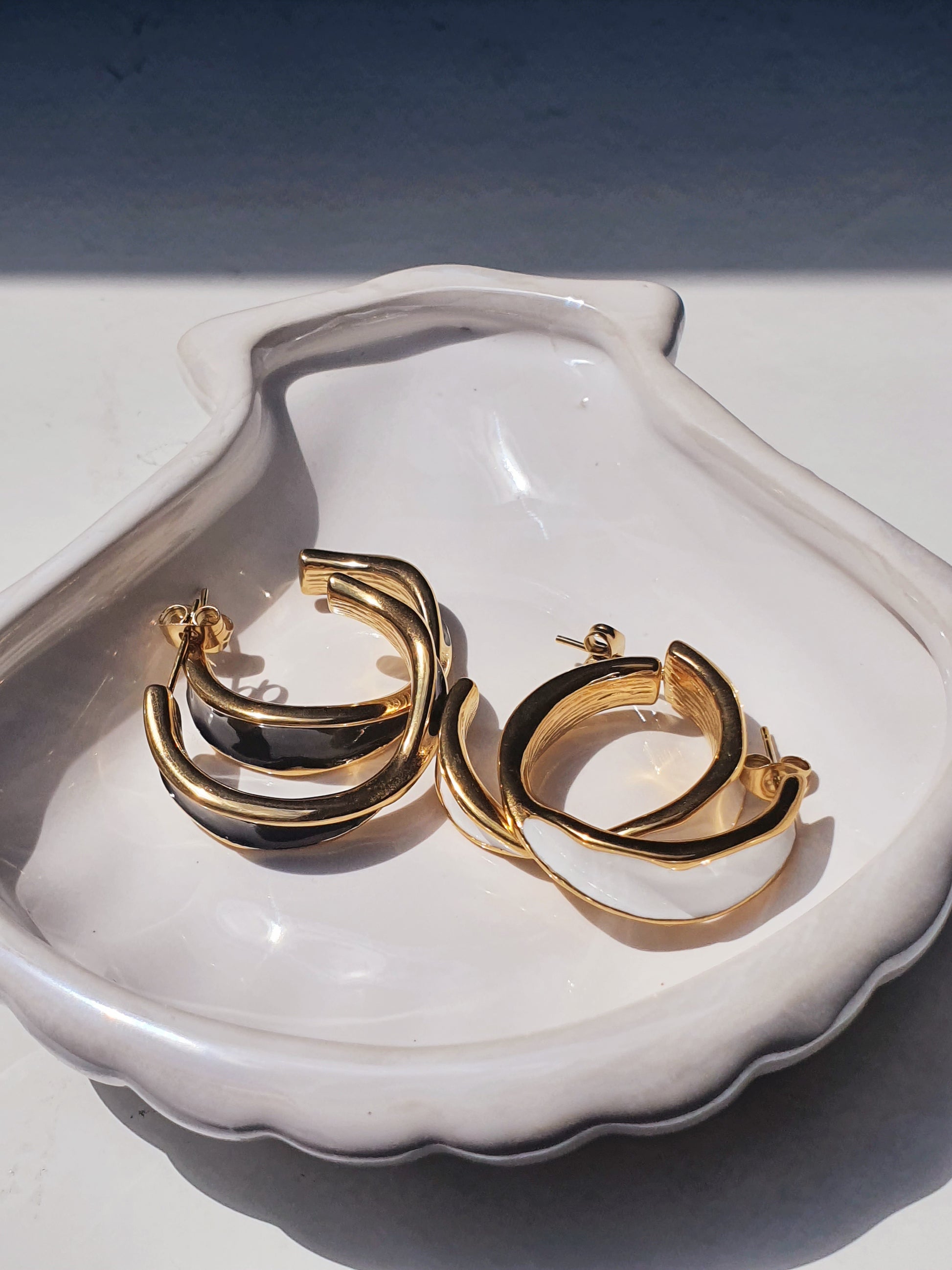 White shell shaped dish holding two pairs of hoops. Both are gold with butterfly stud backs and a twist design. One pair has a white enamel finish and the other pair has a black enamel finish