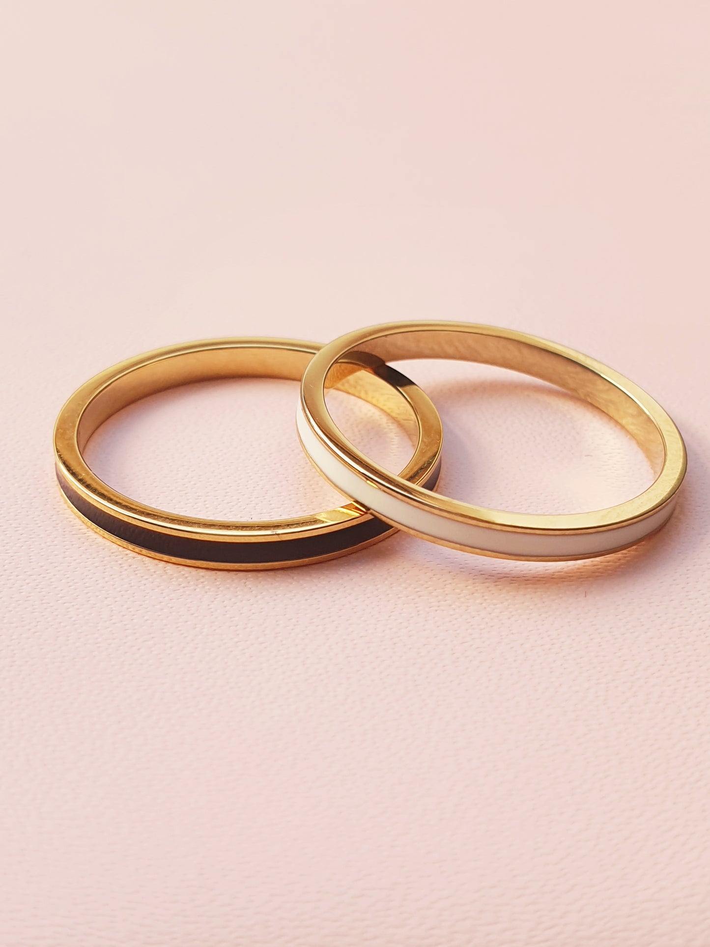 A gold band ring with white enamel banding is stacked on top of another ring with black banding 