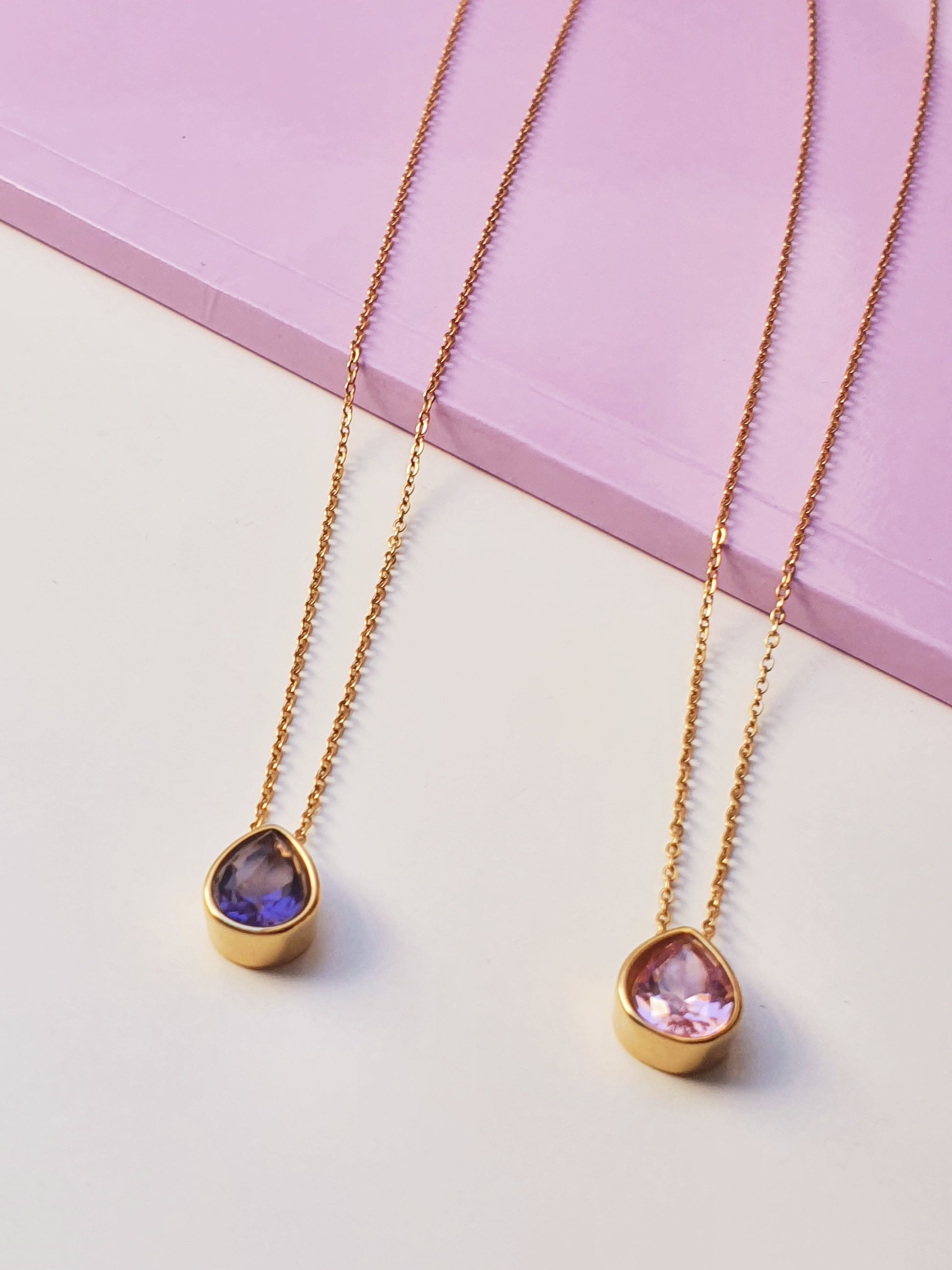 Two gold necklaces with teardrop pendants reast on a purple book and white surface. One pendant has a purple stone and the other has a pale pink stone 