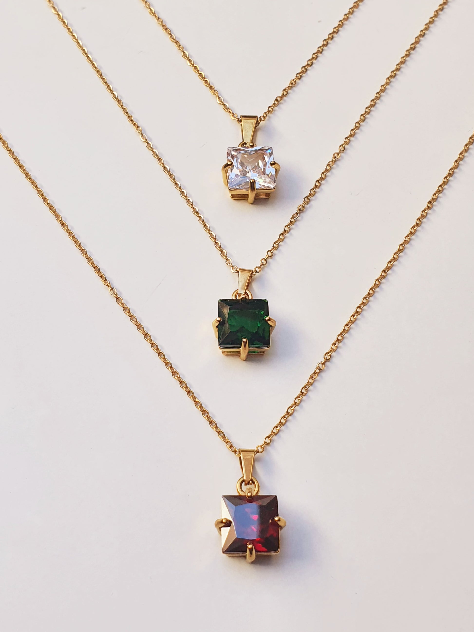 Three gold necklaces with square crystal pendants laid flat. One crystal is clear, another is  a deep green and the last is red