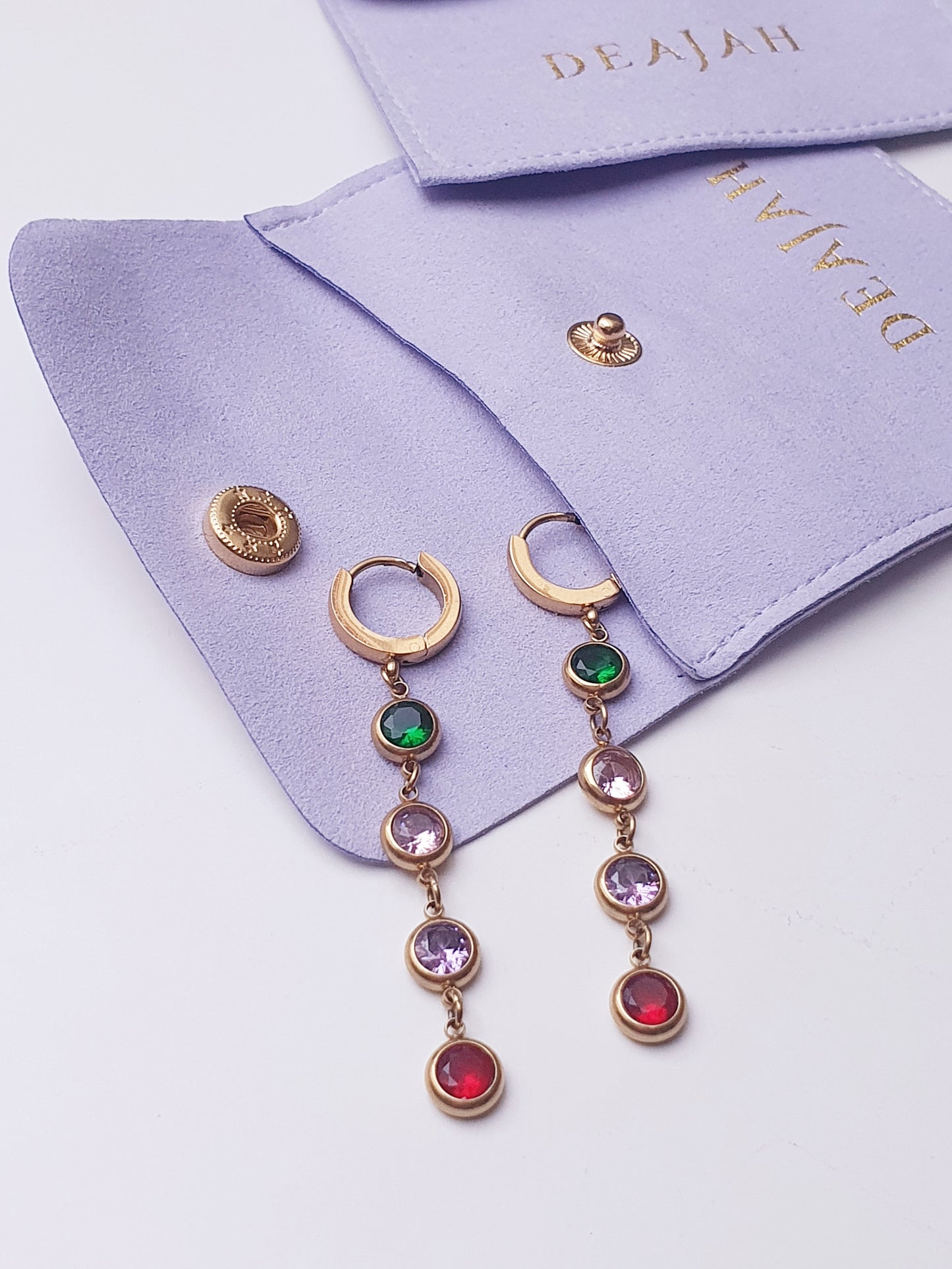 A pair of gold huggy hoops with dangling round gems set with green, pink, purple and red stones rest on a purple jewellery pouch
