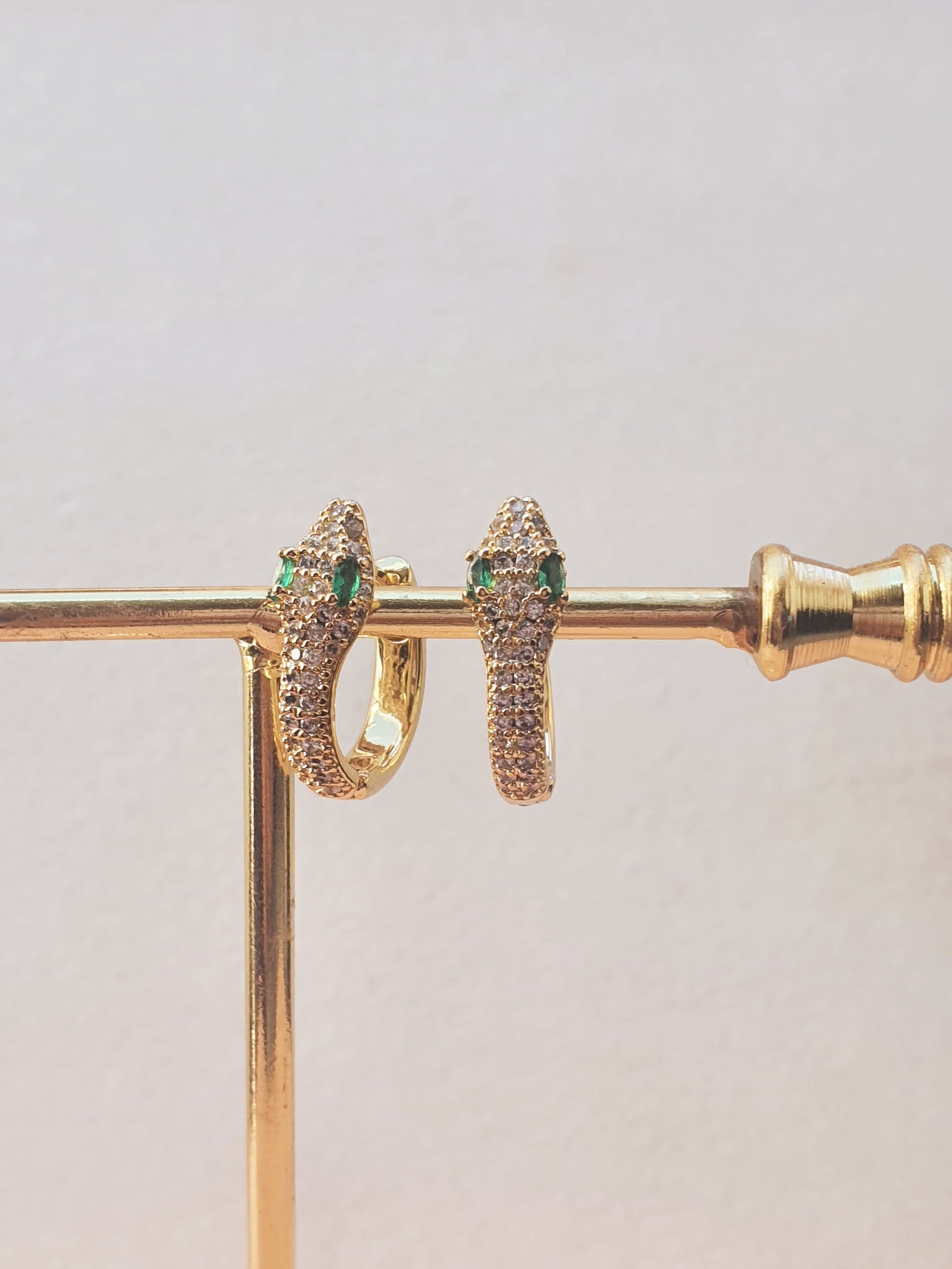 A pair of gold snake shaped huggies set with clear stones and green gems for eyes