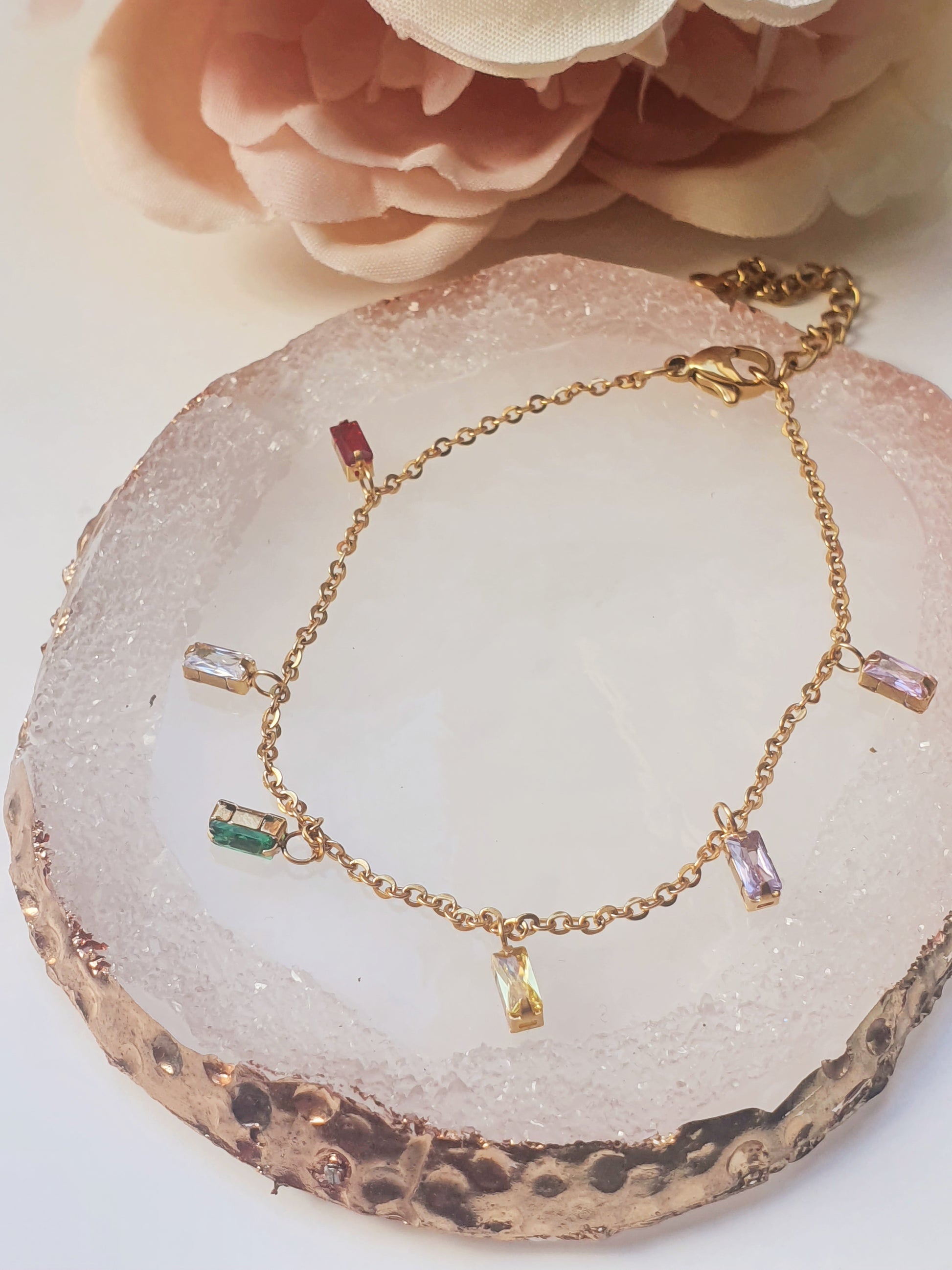 A dainty gold bracelet with dangling gems in multiple colours sits on a clear agate coaster