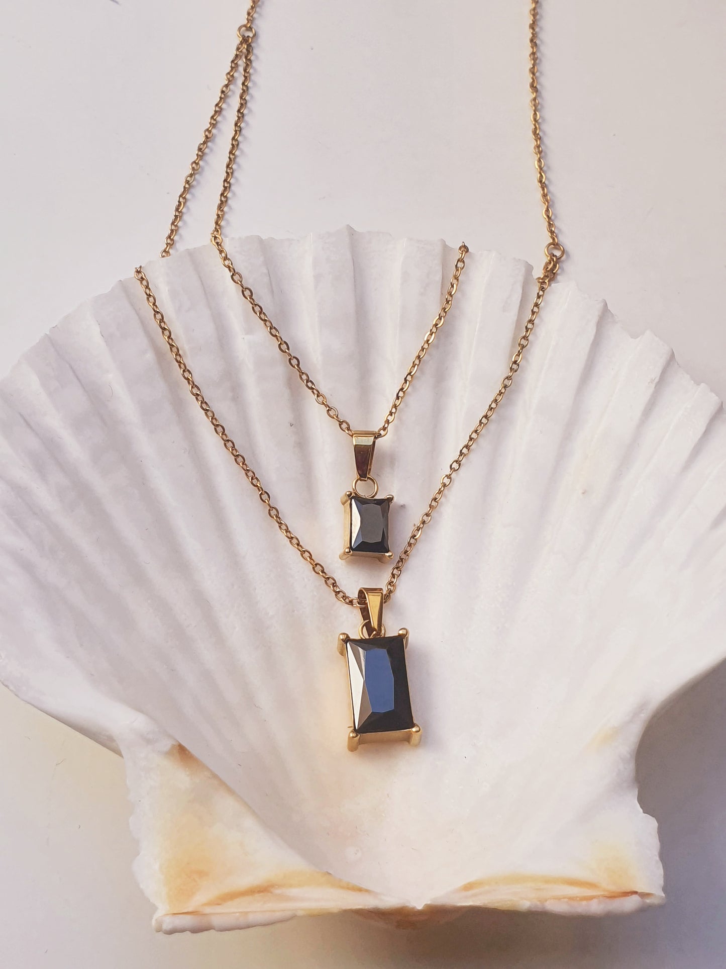 A gold double layered necklace with two black rectangle jewel pendants rests on a large white shell