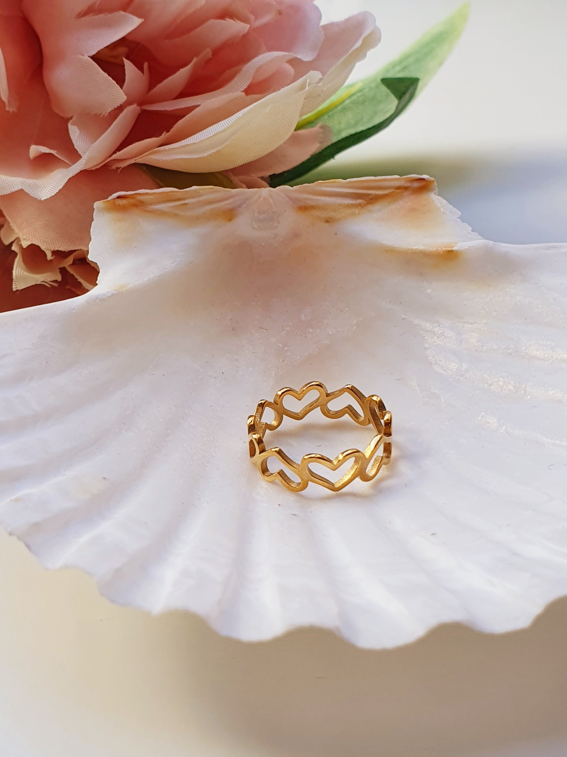 A gold ring made up of connected stencil hearts sits on a large white sea shell with a  pink flower in the background