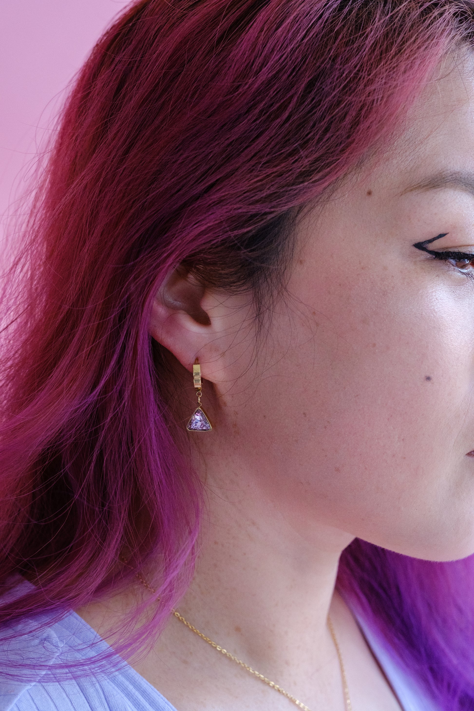 Side profile of a woman's face wearing a gold dangle huggy hoop with triangle pendant set with a pale pink stone