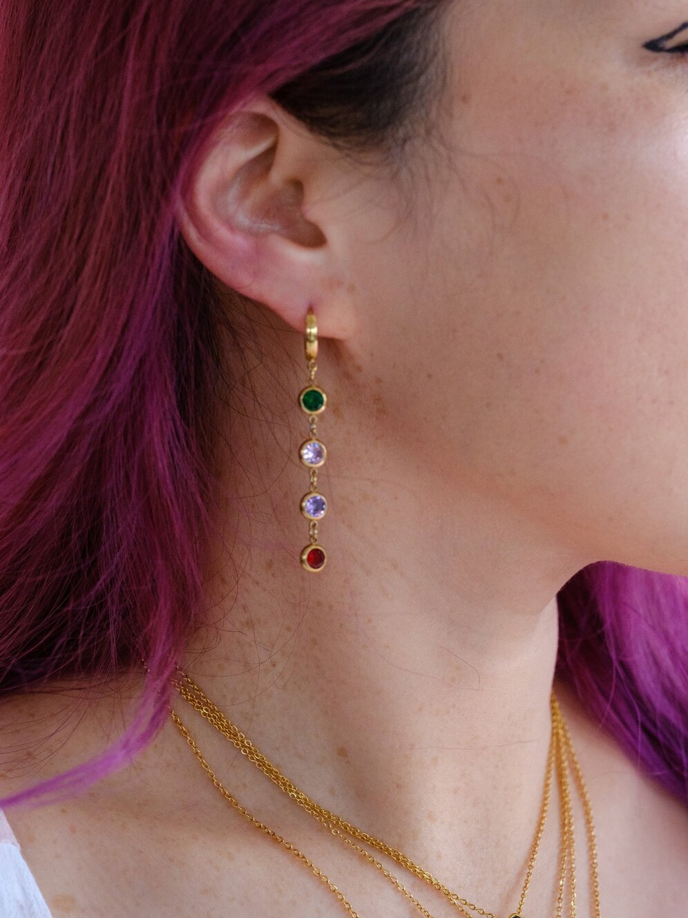 A woman wears a huggy hoop with dangling round gems set with green, pink, purple and red stones