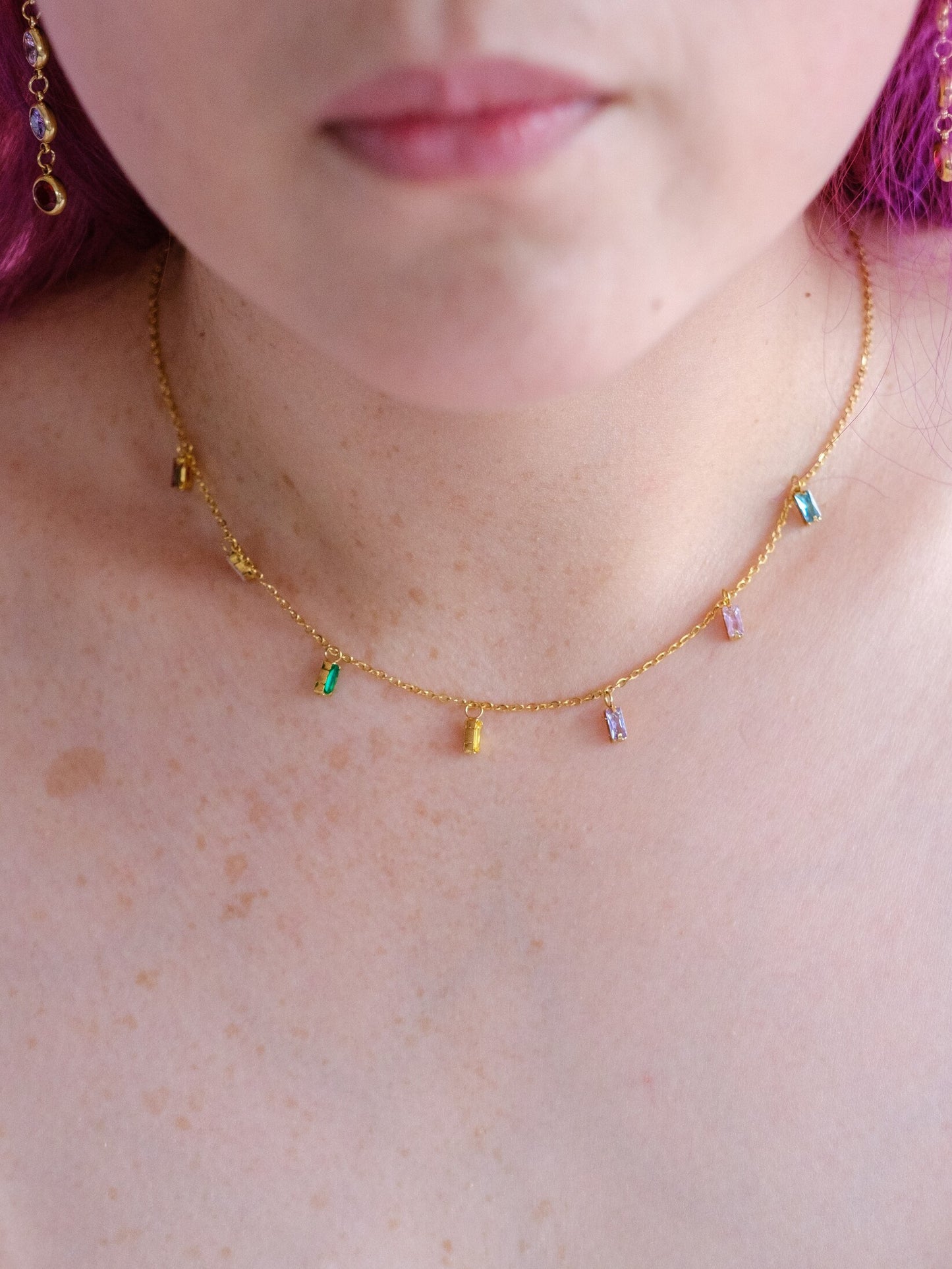 A woman wears a dainty gold necklace with dangling gems in multiple colours