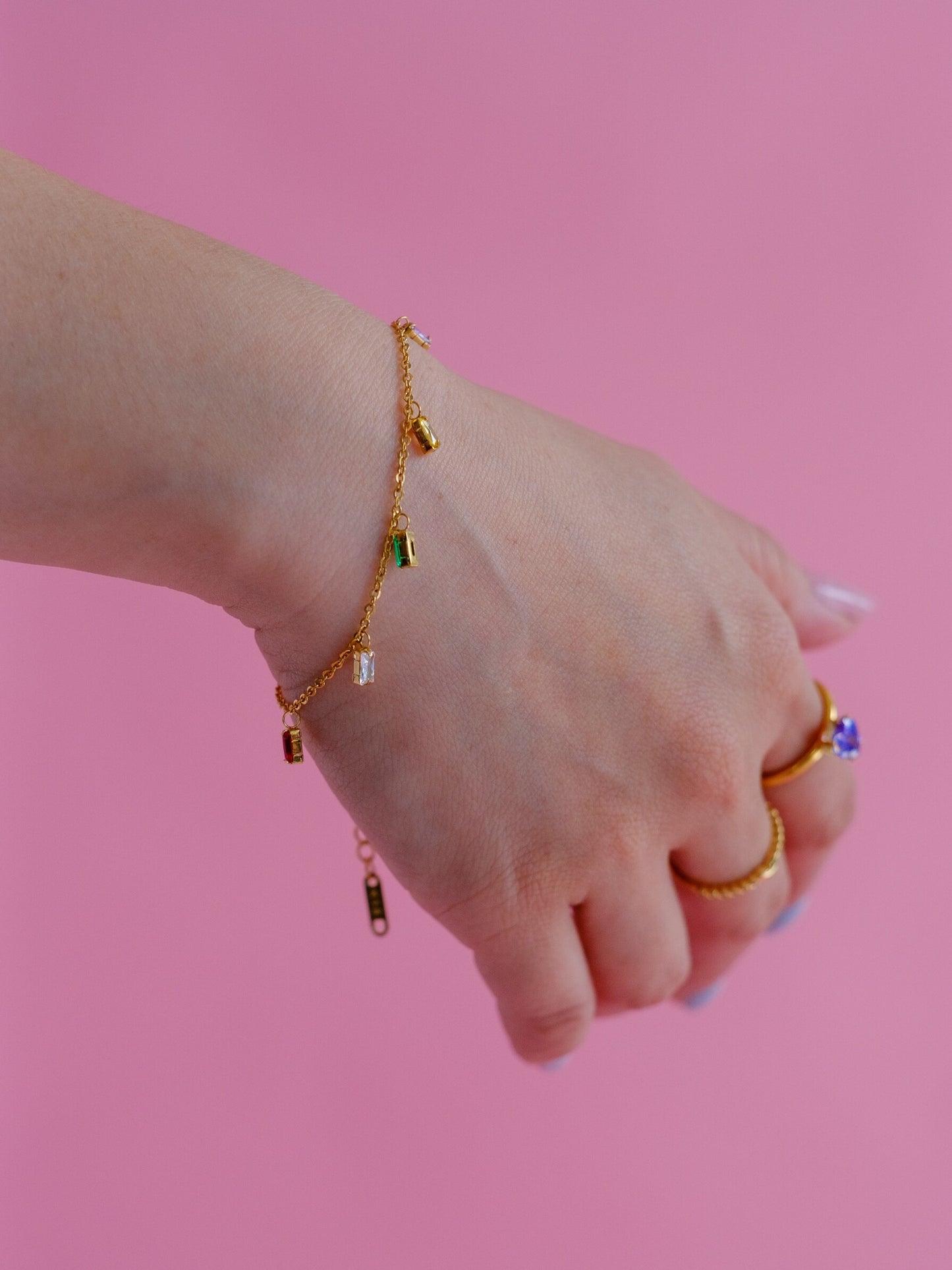 A woman wears a dainty gold bracelet with dangling gems in multiple colours