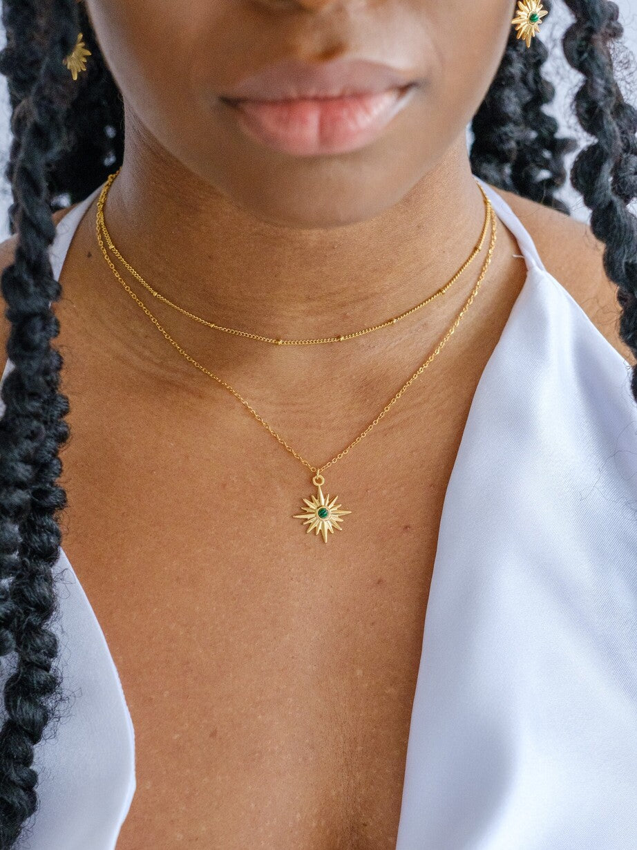 Woman wears two gold necklaces. The shortest necklace is a satellite necklace and the longer necklace has a star shaped pendant set in the centre with a green gem