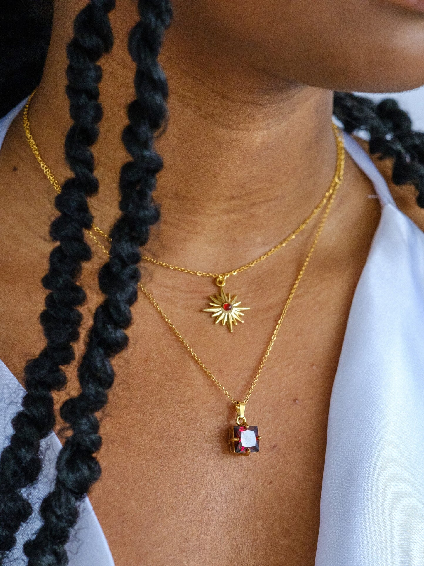 A woman wears two gold necklaces. The shorter necklace has a star shaped pendant with a small red gem in the centre. The longer necklace has a square shaped red crystal pendant