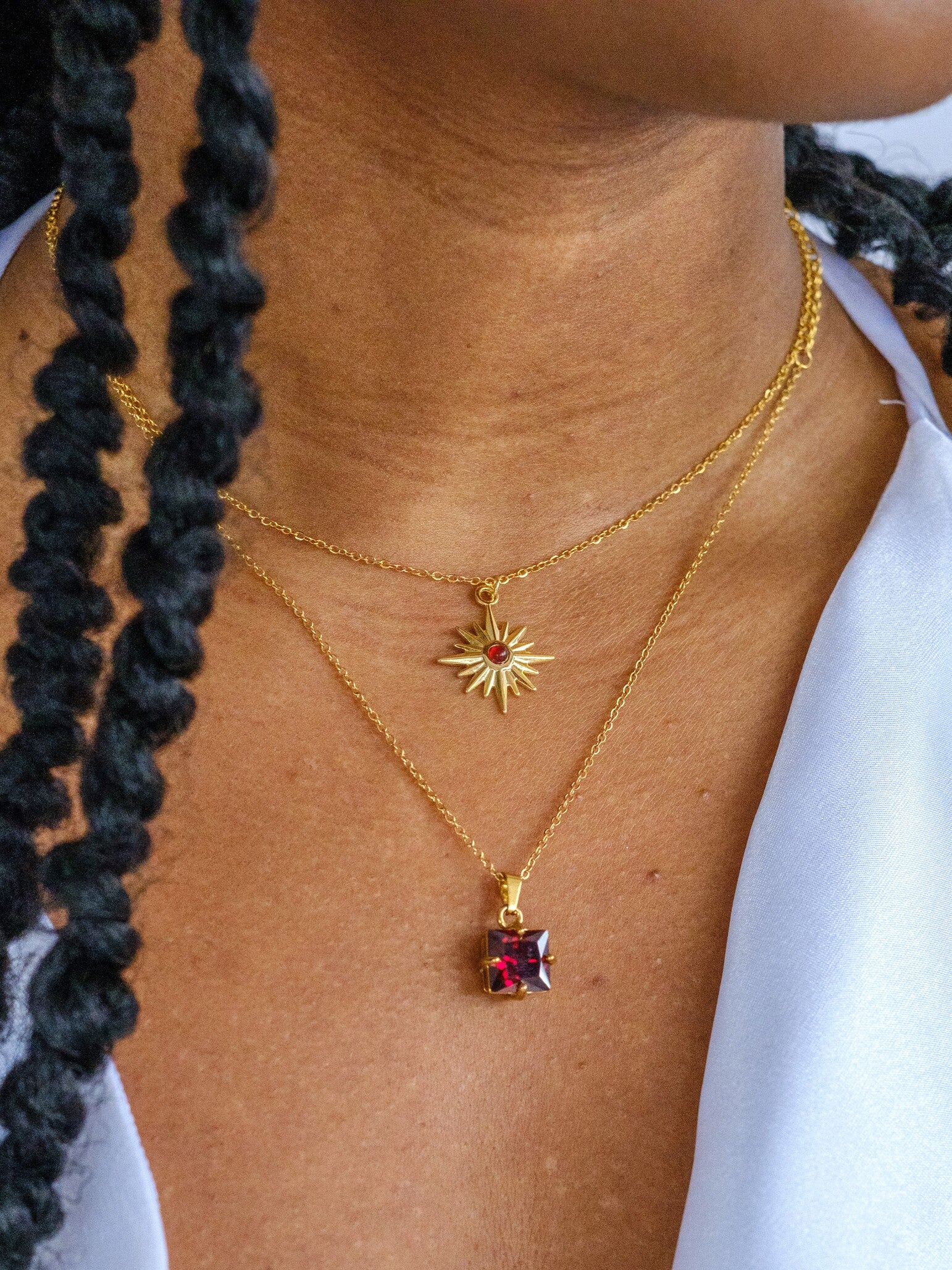 Woman wearing two gold necklaces, one short necklace with a star shaped pendant and a red central gem. The second longer necklace has a red square crystal pendant
