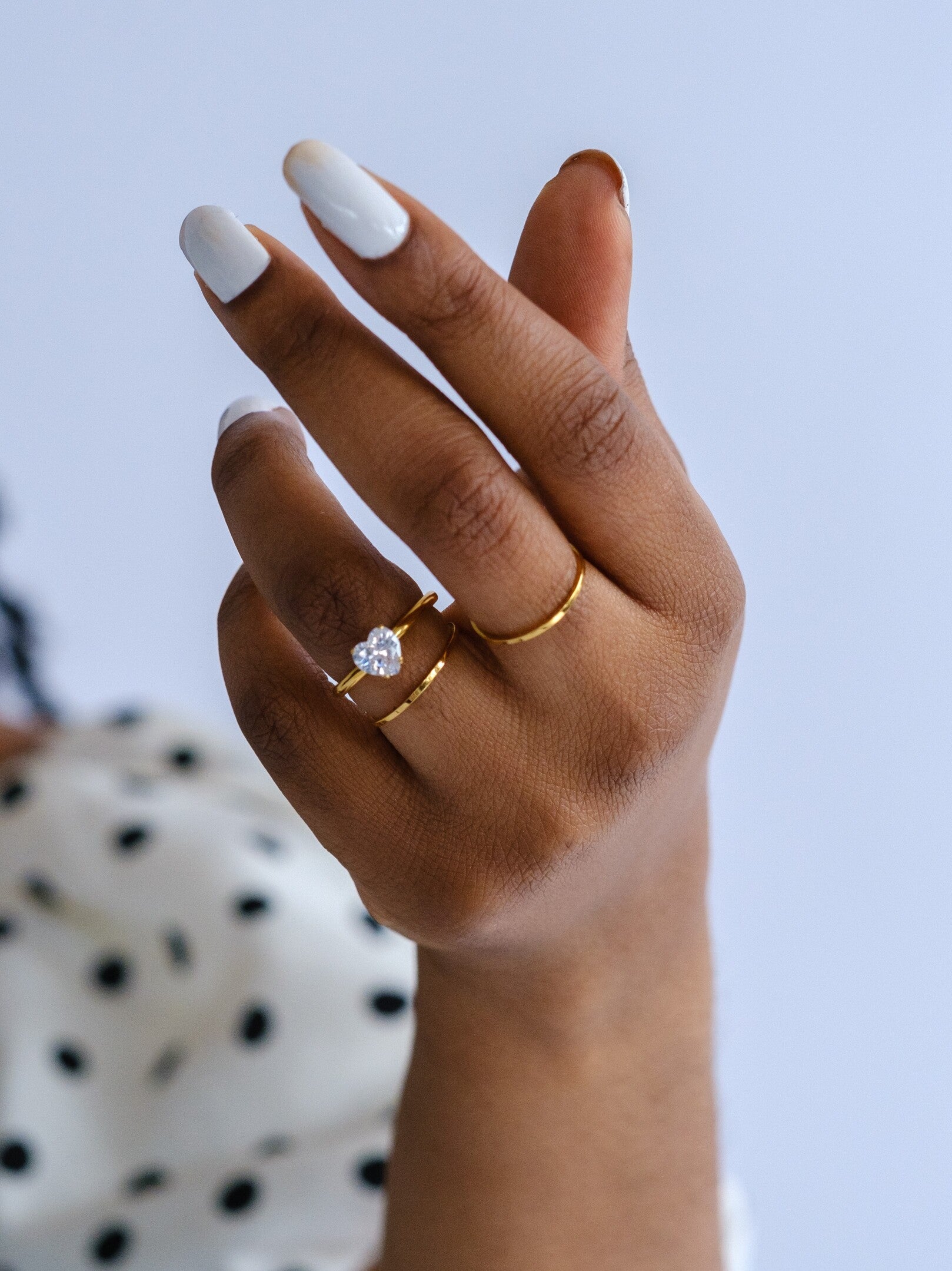 Womans' hand wearing 3 gold band rings. One ring has a clear heart crystal, the other two are slim stacking rings