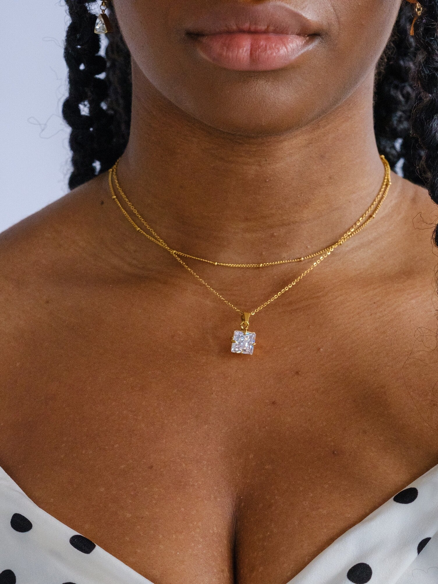 Woman wearing two necklaces. One is a gold satellite necklace at choker length and the other has a clear square crystal pendant and sits a bit lower