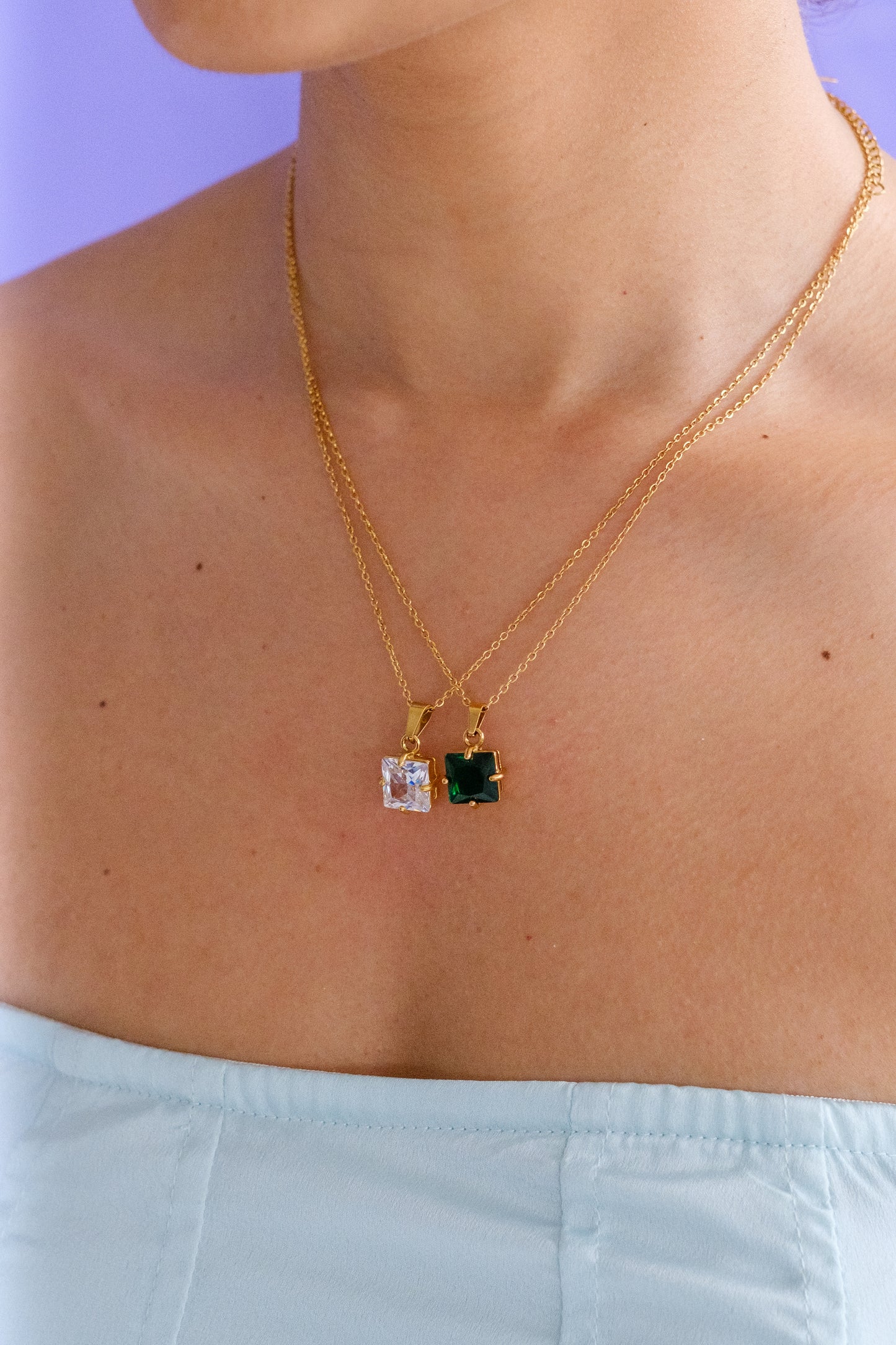Woman wearing two matching gold necklaces. Both with square pendant crystals; one with a green gem and the other with a clear gem