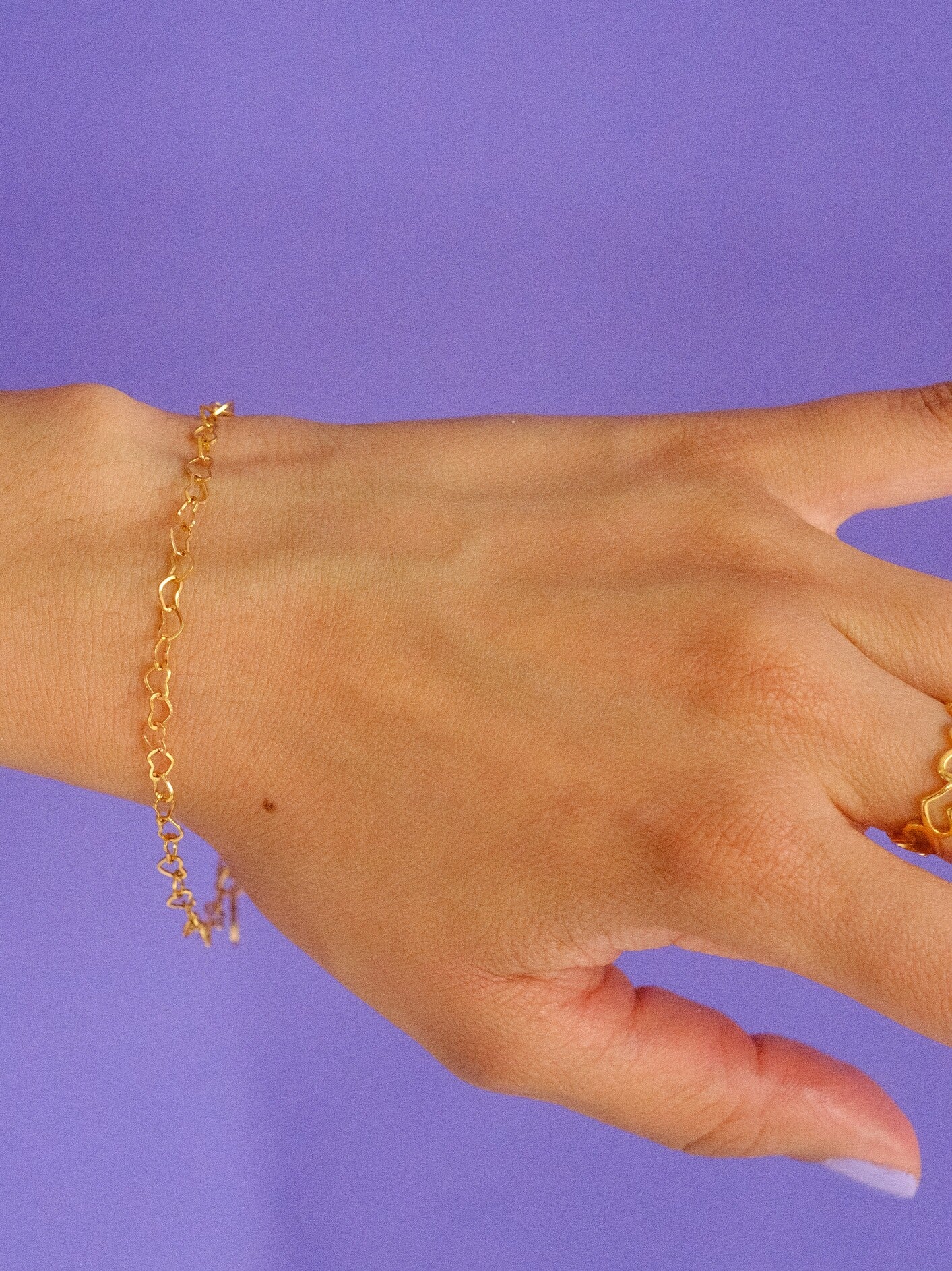 Woman's wrist against a purple background, wearing a gold bracelet with heart shaped links