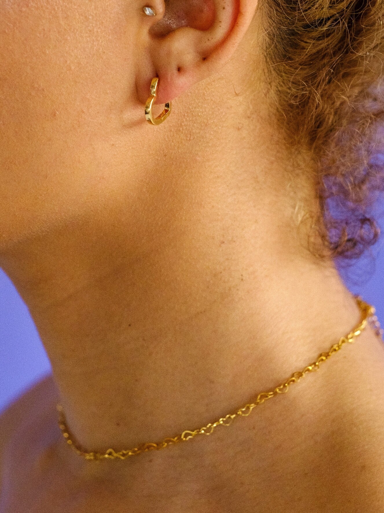 Woman wearing a gold choker made up of heart shaped chain links. Necklace is paired with a gold heart shaped huggy earring