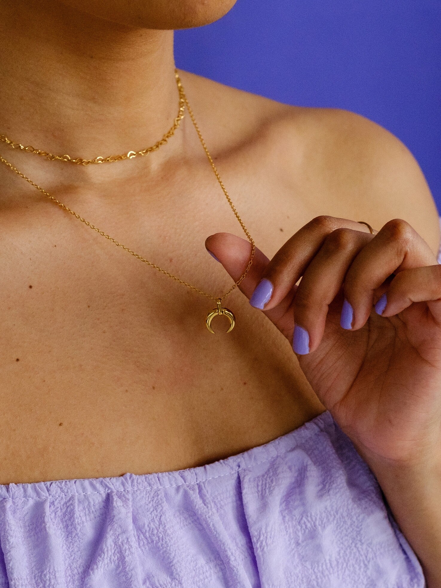 A woman wears two gold necklaces. One necklace is a choker made up of heart shaped chain links. She hold the other necklace which has a crescent moon pendant
