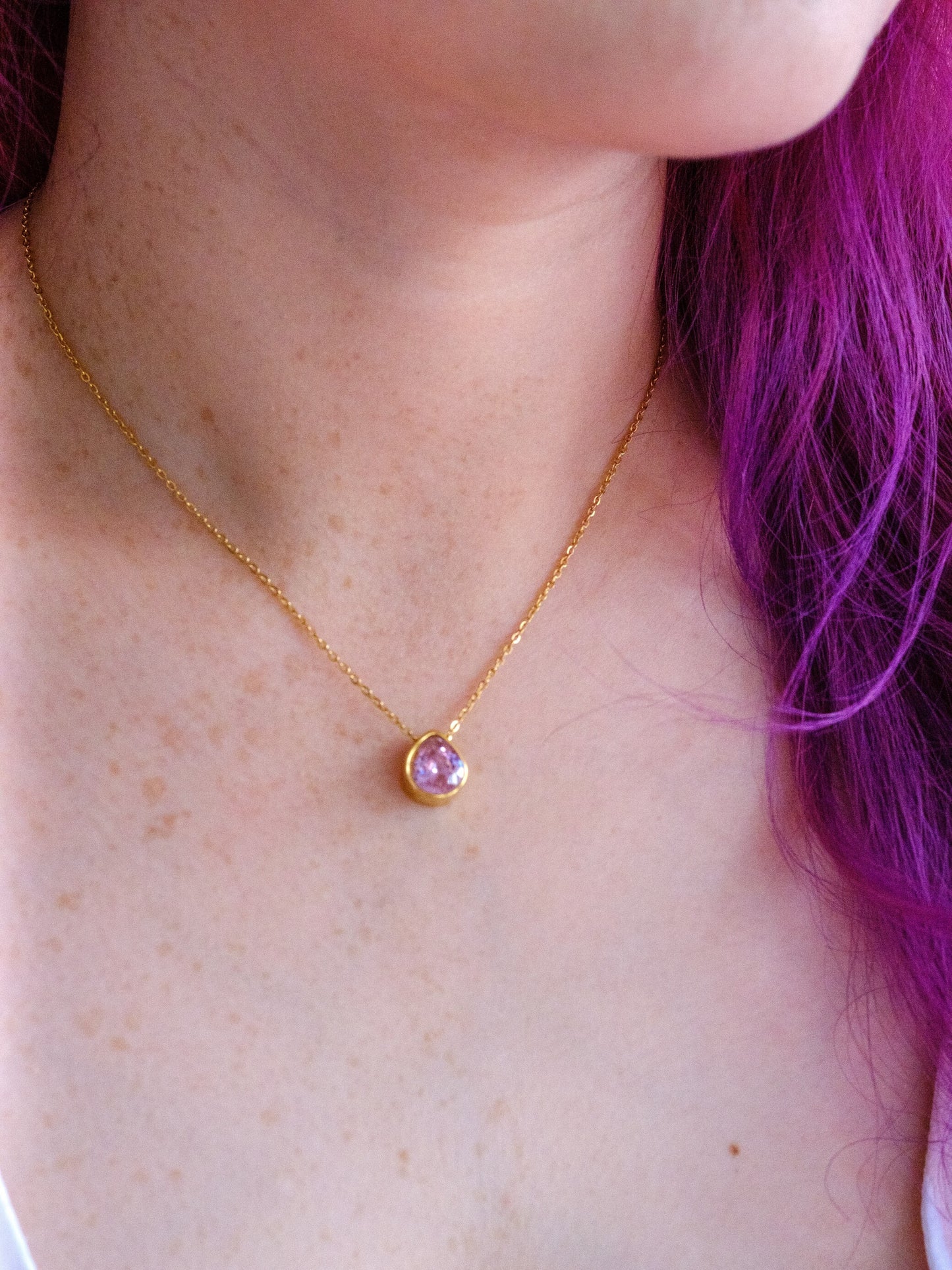 A woman with bright purple hair wears a gold necklace with a teardrop pendant set with a pale pink stone