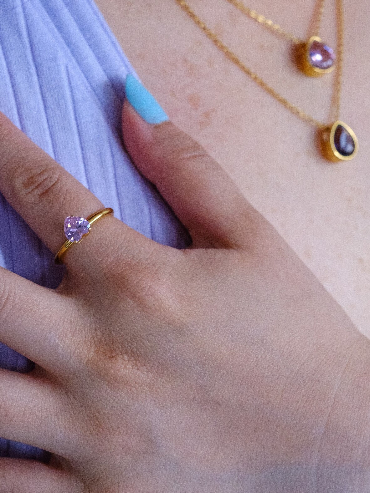 Woman's hand wearing a gold band ring with a pale pink heart gem stone. The woman also wears two gold necklaces with tear drop pendants with a pink and a purple gem