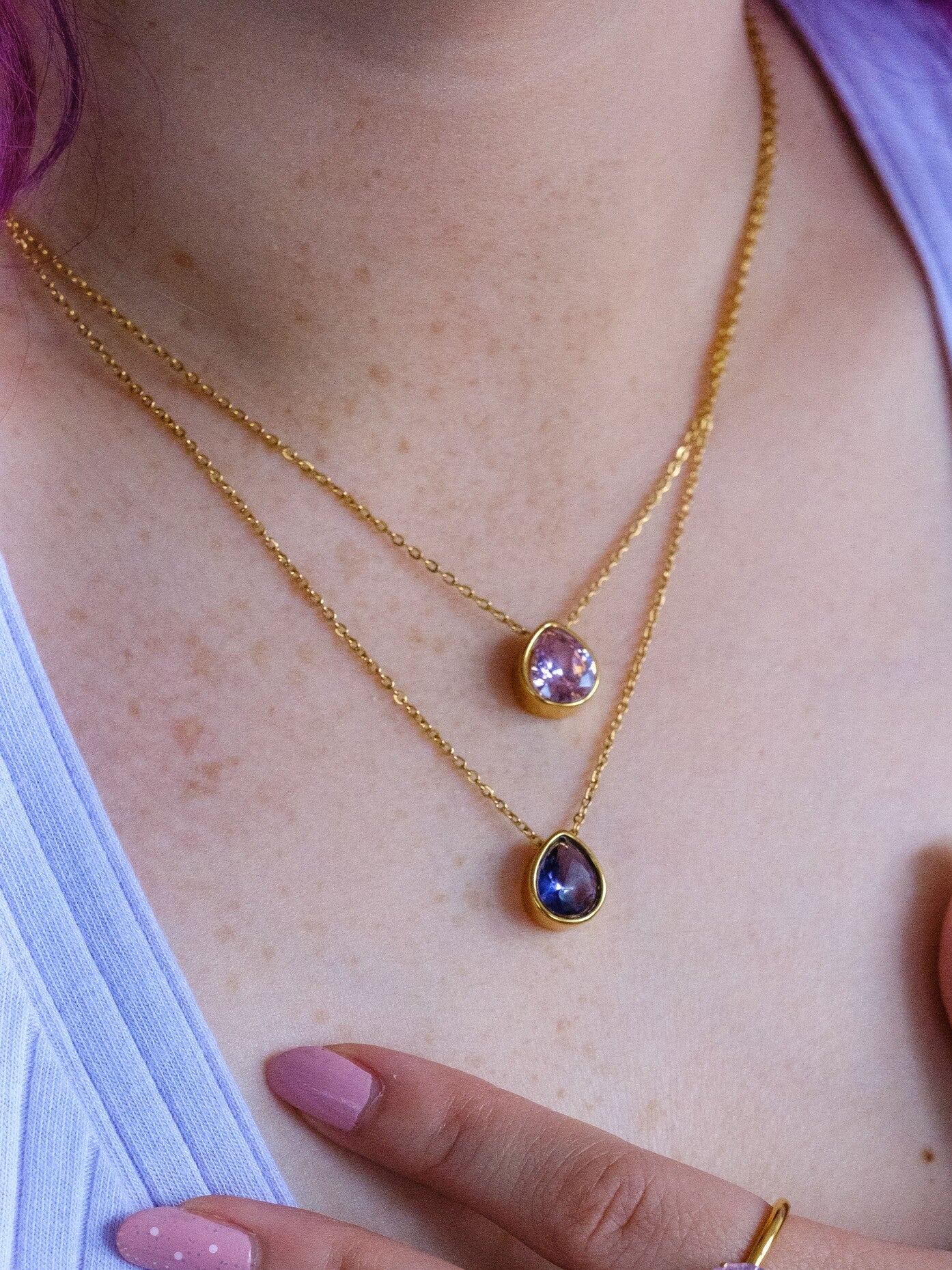 A woman wears to gold necklaces with teardrop pendants. The top pendant is set with a pale pink stone and the lower necklace is set with a purple stone