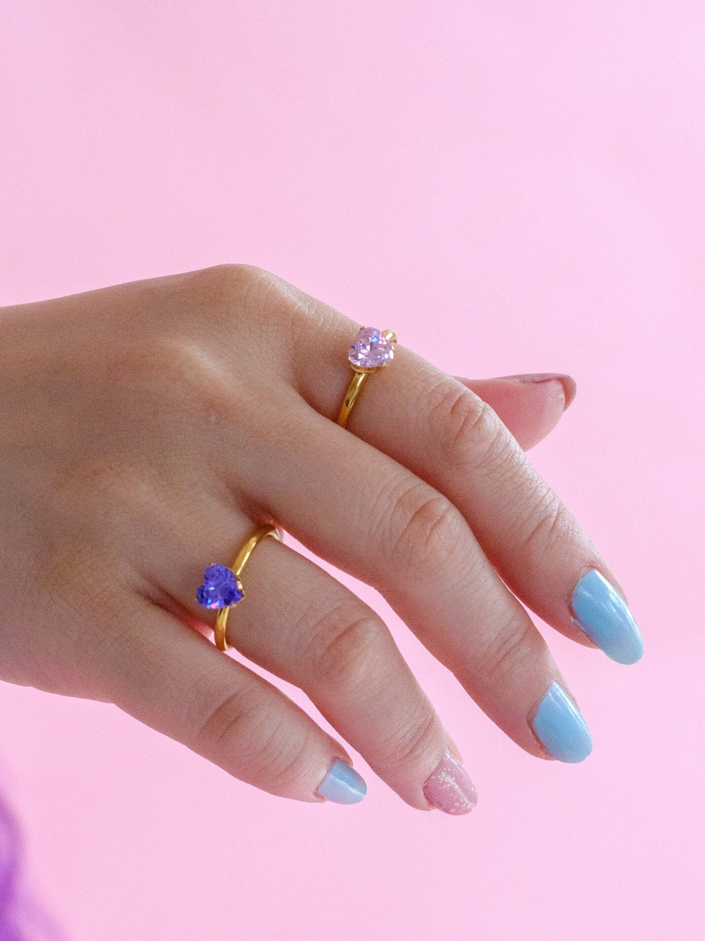 Woman's hand wearing two gold band rings. One ring has a purple heart shaped gen and the other has a pale pink heart shaped gem
