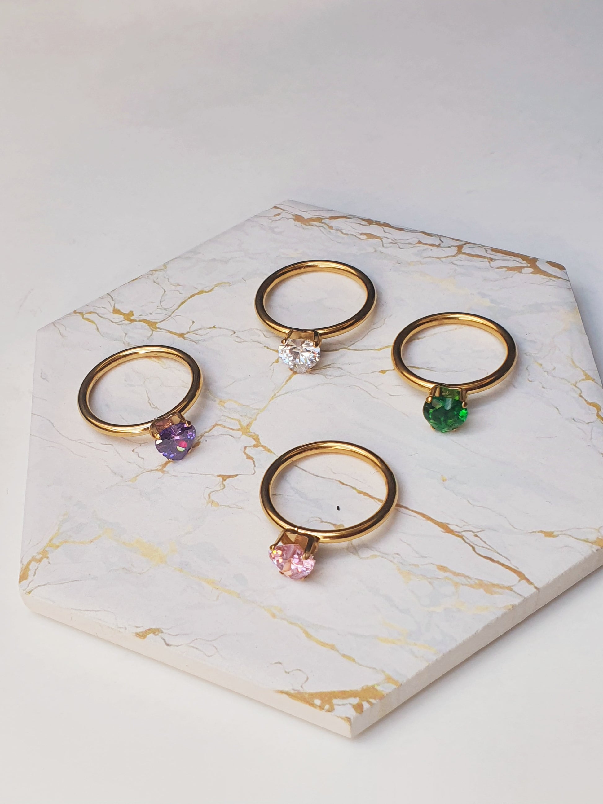 Four gold band rings on a marble coaster. All have different colour heart gems in purple, pink, clear and green crystal 