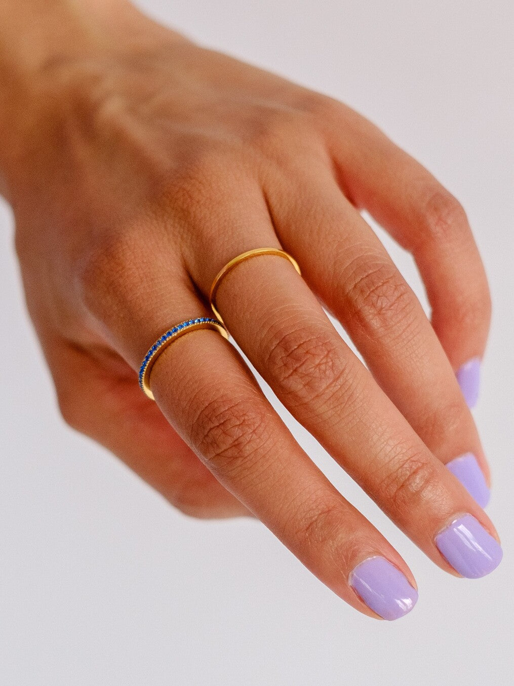 Woman's hand with purple nail polish wearing two gold rings. One ring is an eternity band set all the way around. The other is a slim gold stacking ring 