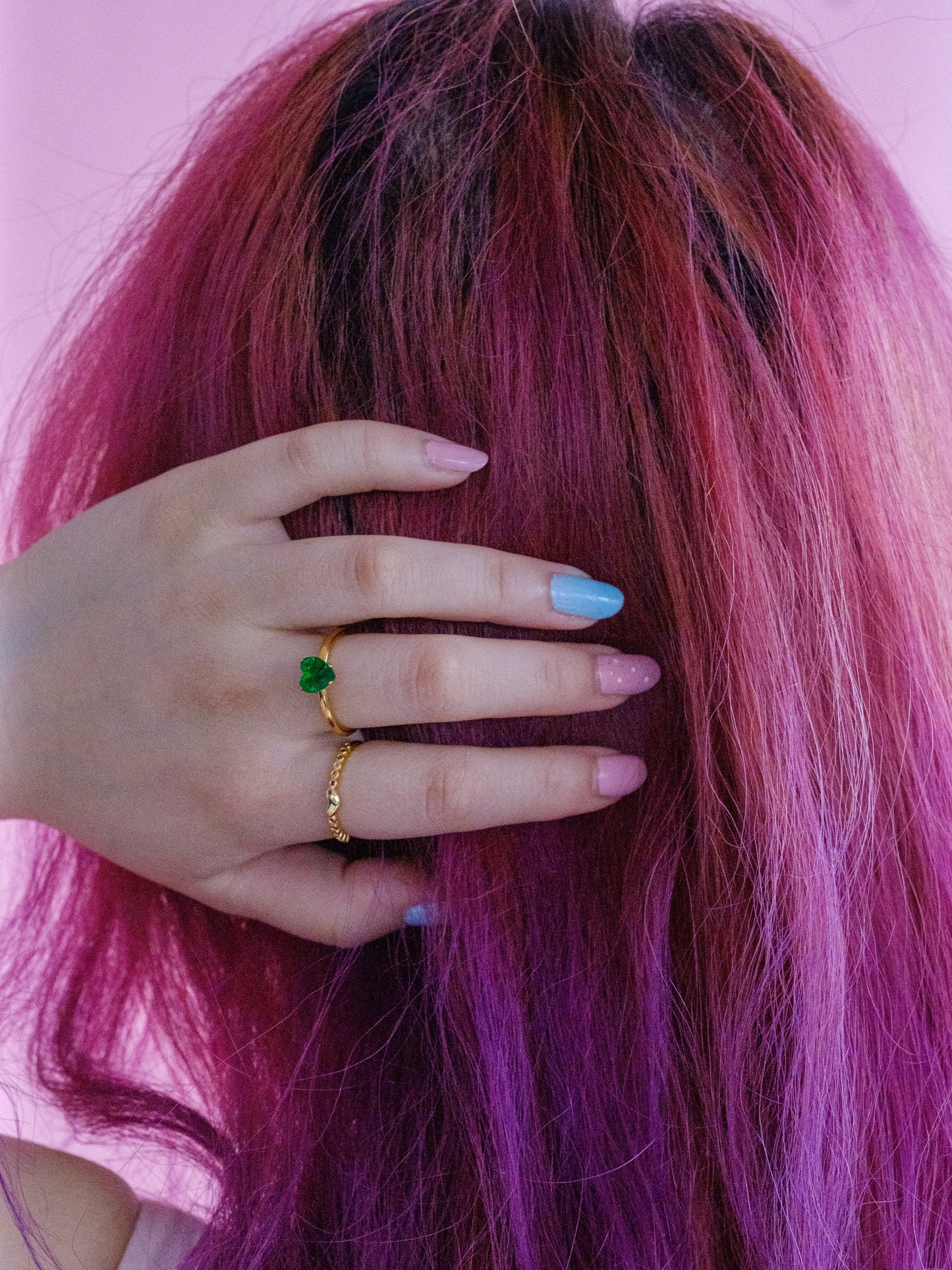 Woman's hand on the back of her head, wearing two gold rings. One with a small gold heart centre and the other with a green heart gem stone
